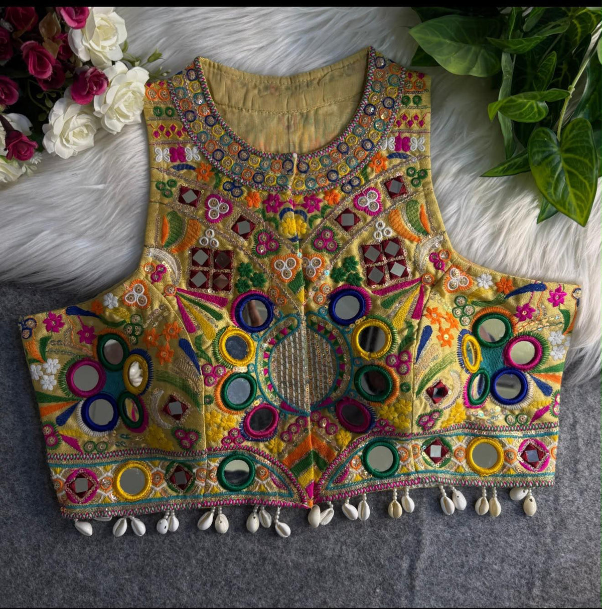 Front view of mustard gold Kutchi halter crop blouse with heavy Rabari mirror embroidery, multicolour floral motifs and cowrie shell fringe hem